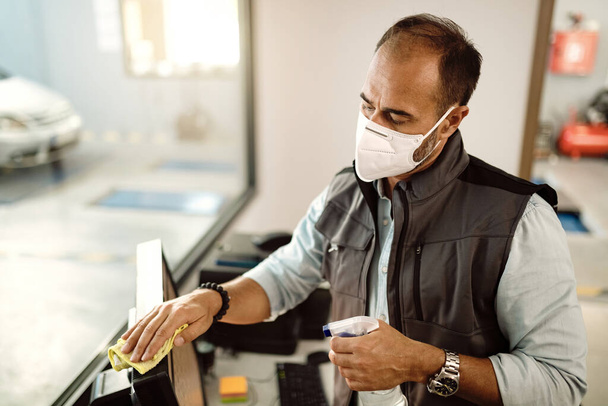 Auto mechanic cleaning office computer with disinfectant and wearing face mask due to COVID-19 pandemic. - Foto, Imagem