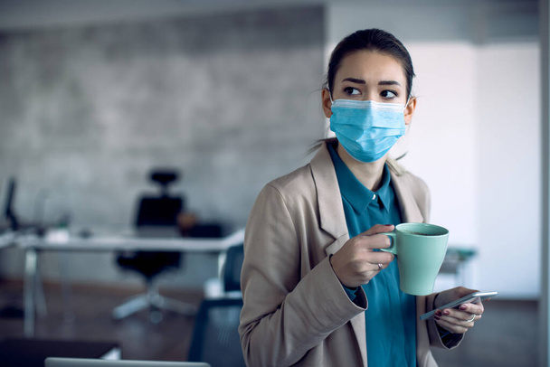 Young businesswoman having a cup of coffee while texting on mobile phone in the office. She is wearing face mask due to COVID-19 pandemic. - Fotografie, Obrázek