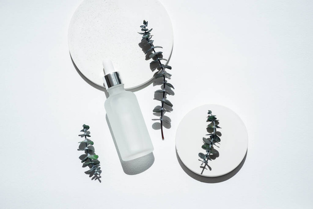 Transparent frosted glass dropper bottle with eucalyptus branches. White background with daylight. Skincare serum or essential oil natural cosmetic. Beauty concept for face and body care - Photo, Image