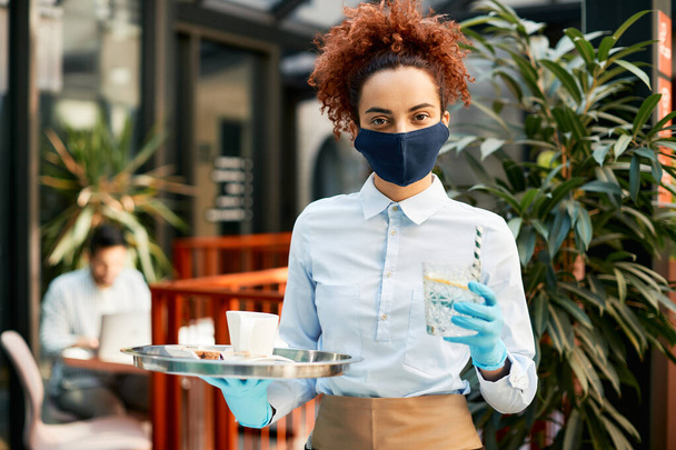 Σερβιτόρα που φοράει μάσκα προσώπου ενώ σερβίρει ποτά σε καφέ που ξανάνοιξε κατά τη διάρκεια πανδημίας του Coronavirus. - Φωτογραφία, εικόνα