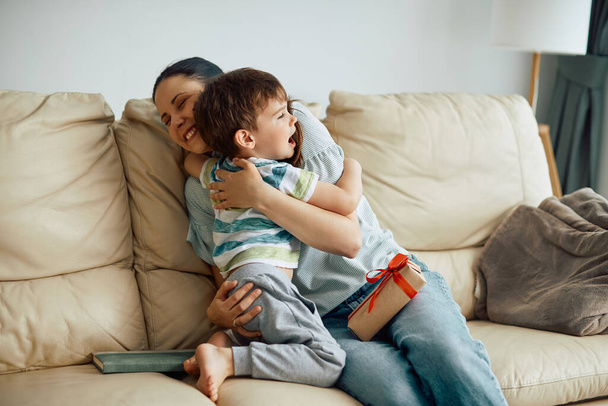 Affectionate little boy embracing his mother while giving her present on Mother's Day at home. - Φωτογραφία, εικόνα