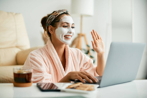 Young happy woman using laptop and waving to someone during video call while wearing white cosmetic face mask. - Fotografie, Obrázek