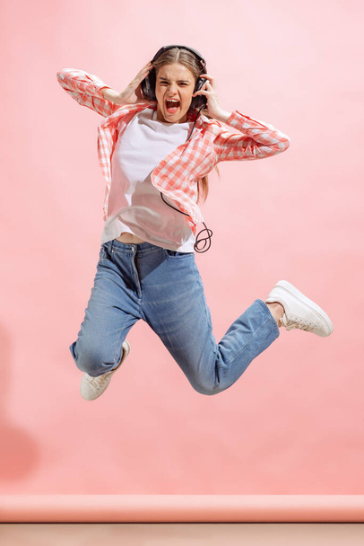 Portrait of excited young girl in checkered shirt listening to music in headphones and jumping isolated over pink studio background. Concept of youth, fashion, lifestyle, emotions, facial expression. - Фото, зображення