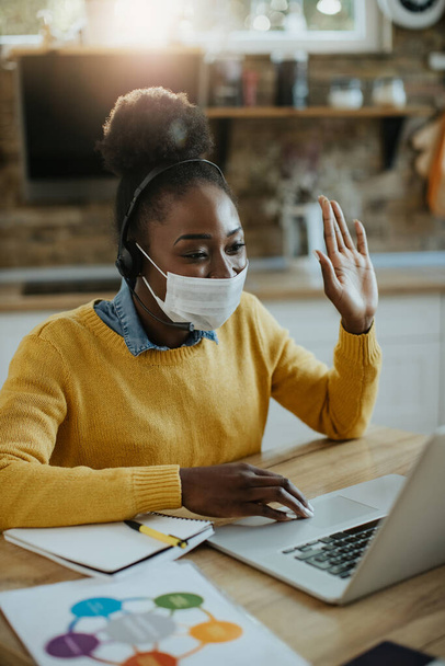 Happy black businesswoman wearing protective face mask and greeting someone during video call over laptop at home.  - Foto, imagen