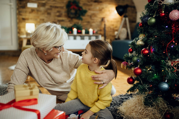 Glückliches kleines Mädchen und ihre Großmutter reden, während sie zu Hause am Weihnachtsbaum sitzen.  - Foto, Bild
