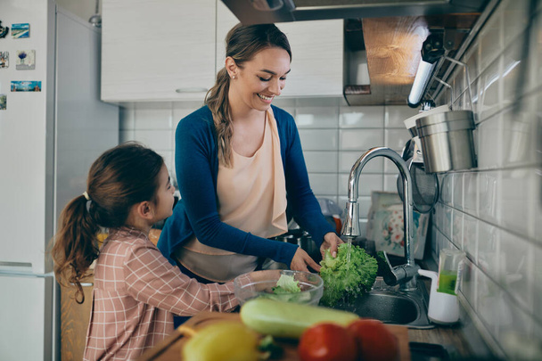 Happy mother and her small daughter washing vegetables under kitchen sink while preparing food together at home.  - Photo, image