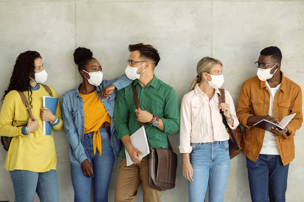 Багатоетнічна група друзів коледжу спілкуються, одягаючи захисні маски для обличчя, стоячи біля стіни у вестибюлі
.  - Фото, зображення