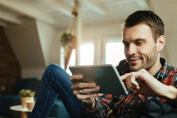 Junger glücklicher Mann liest etwas auf Touchpad und entspannt im Wohnzimmer.  - Foto, Bild