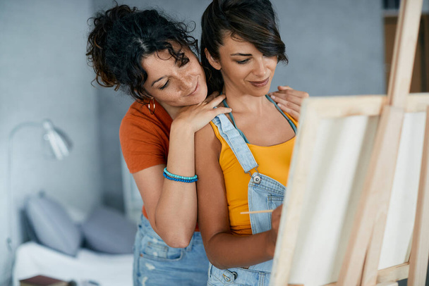 Young lesbian woman painting on canvas while her girlfriend is standing behind her. - Foto, afbeelding