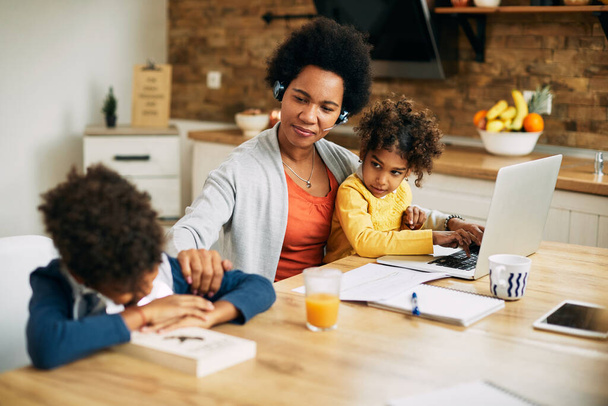 Happy African American stay at home mother talking to her son who is feeling tired of learning at home. Small daughter is sitting on her lap and using laptop.  - 写真・画像