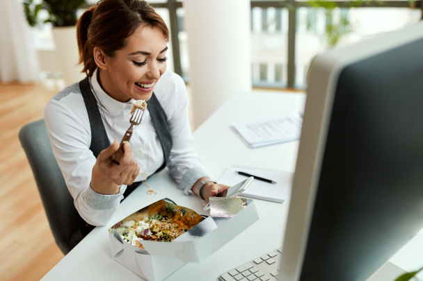 Young smiling businesswoman texting on cell phone while eating at her office desk.  - Foto, imagen
