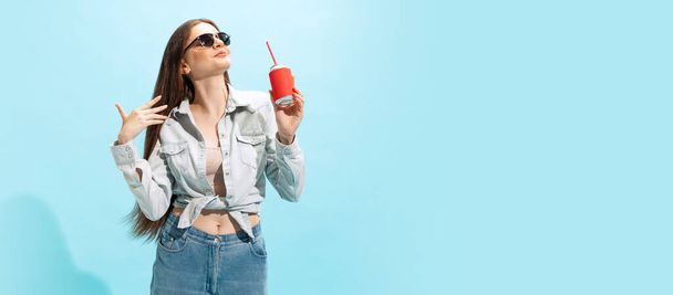 Portrait of stylish young girl in casual clothes and sunglasses posing with soda isolated over blue background. Concept of youth, fashion, lifestyle, emotions, facial expression. Copy space for ad - Фото, изображение
