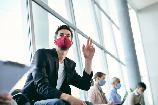 Молодой бизнесмен ждет собеседования и поднимает руку во время переклички. Он носит маску из-за пандемии коронавируса. - Фото, изображение