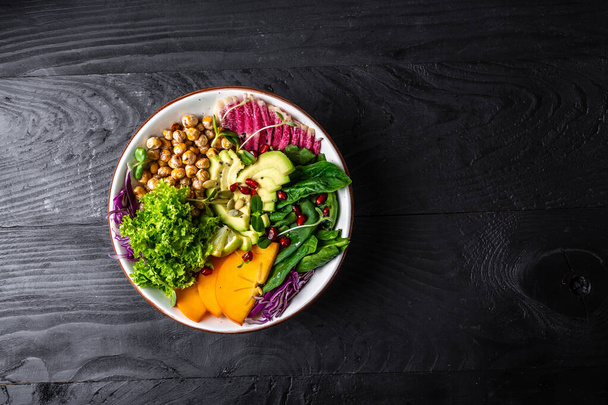 Fresh salad with roasted chickpeas, avocado, persimmon, spinach, avocado, watermelon radish and seeds on a dark background. Long banner format. top view. - Photo, Image