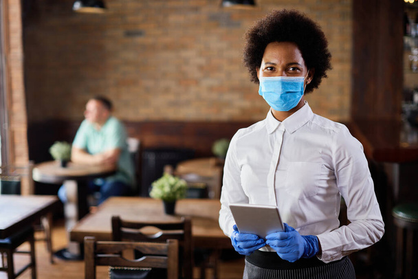 Μαύρη σερβιτόρα που χρησιμοποιεί touchpad ενώ χρησιμοποιεί ψηφιακή ταμπλέτα και φοράει προστατευτική μάσκα προσώπου λόγω πανδημίας του coronavirus.  - Φωτογραφία, εικόνα