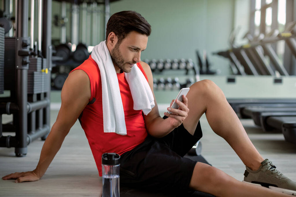 Спортсмен смс на смартфоні, сидячи на підлозі в клубі здоров'я
.  - Фото, зображення