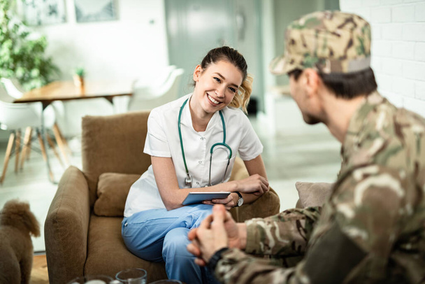 Happy nurse using touchpad and communicating with a soldier while visiting him at home.  - Foto, Imagen