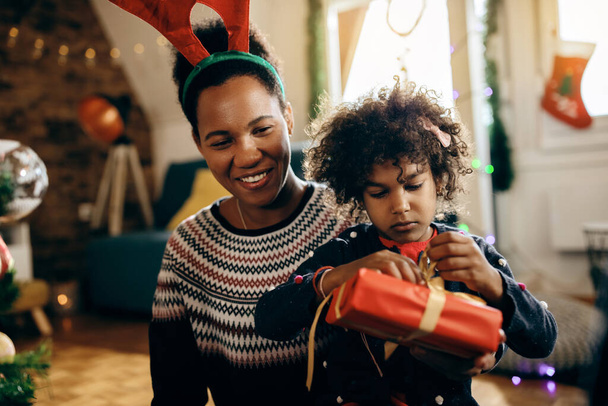 Happy African American mother enjoying with her daughter who is unwrapping Christmas present at home.  - Zdjęcie, obraz