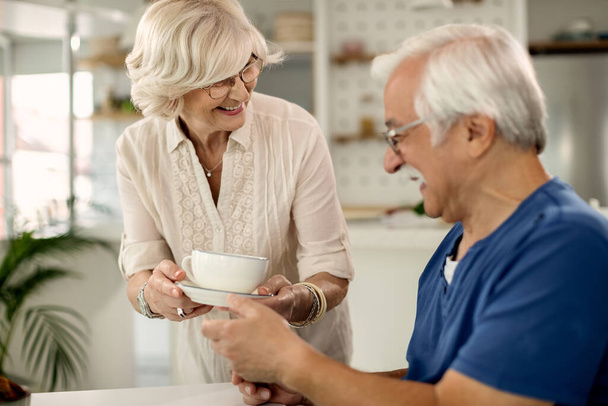 Ευτυχισμένη ηλικιωμένη γυναίκα που δίνει πρωινό καφέ στον άντρα της στο σπίτι..  - Φωτογραφία, εικόνα