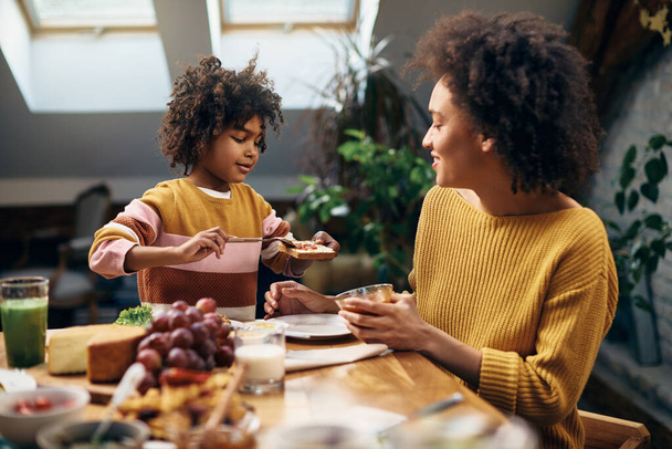Little black girl spreading jam on toasted break while eating breakfast with mother in dining room. - Foto, Imagem