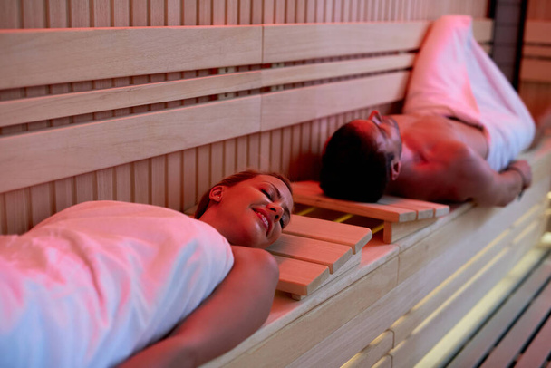 Smiling couple relaxing with eyes closed while lying down in sauna at the spa. Focus is on woman.  - Foto, Imagem