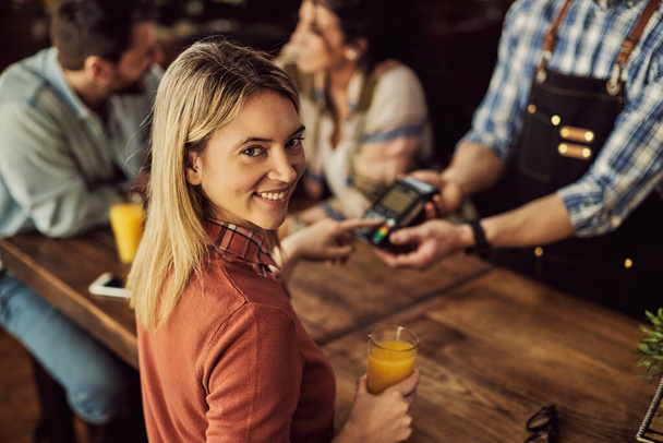Молода щаслива жінка друкує шпильку при оплаті кредитною карткою в кафе і дивиться на камеру
.  - Фото, зображення