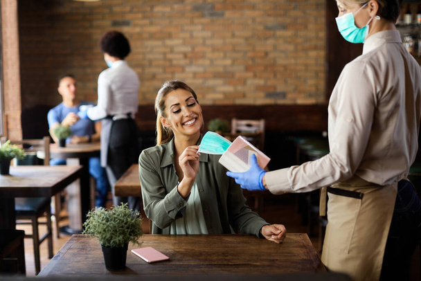 Waitress offering to young woman protective face mask in a cafe during coronavirus epidemic. Focus is on woman.  - Foto, imagen