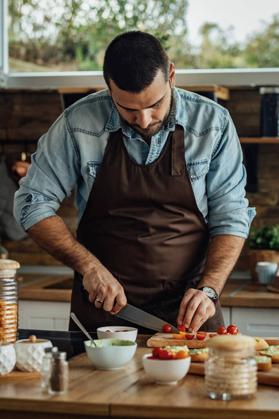 Молодий чоловік ріже вишневий помідор на обробній дошці, готуючи здорову їжу на кухні
.  - Фото, зображення