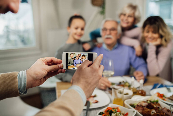 Close-up of man using smart phone while photographing his extended family in dining room. - Foto, afbeelding
