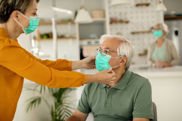 Adult daughter adjusting senior father's protective face mask at home during virus pandemic. Focus is on man.  - Foto, Imagem
