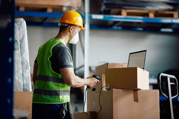 Warehouse worker with face mask using laptop and scanning packages at storage compartment. - Foto, imagen