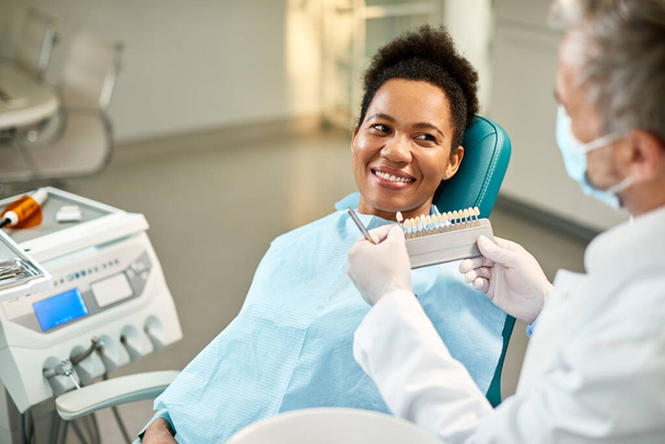 Happy African American woman and her dentist choosing the right shade of implants during dental appointment. - Foto, Bild