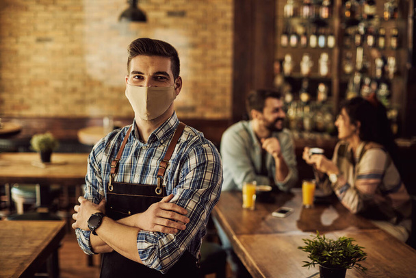 Portrait of confident waiter with protective face mask standing with crossed arms in a cafe. There are people in the background.  - Foto, Bild