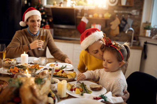 Milosrdná matka líbání své malé dcery, zatímco mají vánoční oběd doma. - Fotografie, Obrázek