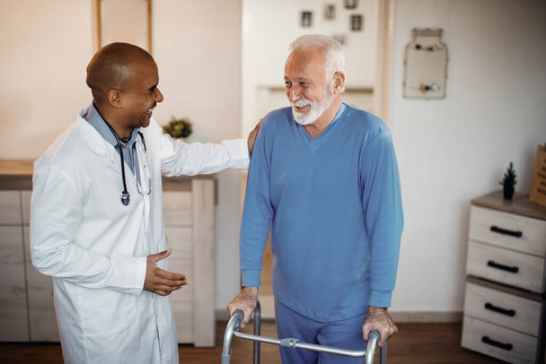 Happy senior man talking to African American doctor while using mobility walker at nursing home.  - 写真・画像
