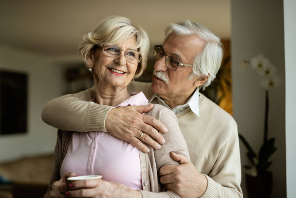 Uśmiechnięty starszy mężczyzna obejmujący swoją żonę podczas rozmowy z nią w domu.  - Zdjęcie, obraz