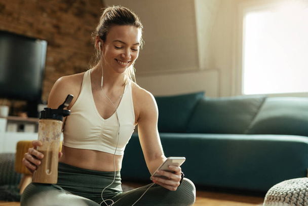 Onnellinen urheilija käyttää älypuhelinta juodessaan proteiinipirtelöä kotona harjoittelun jälkeen.  - Valokuva, kuva