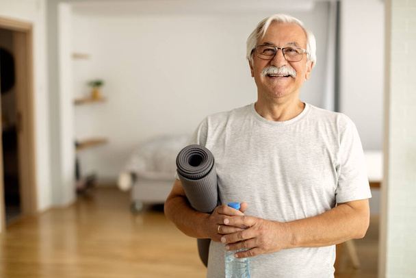 Happy senior man holding bottle of water and exercise mat in the living room and looking at camera.  - 写真・画像