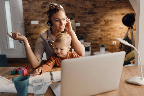 Distraught working mother talking over headset while taking care of her small son at home.  - Valokuva, kuva