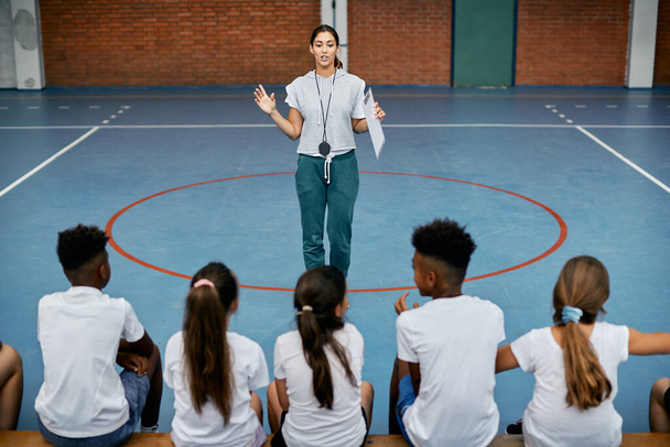 Young female coach talking to group of elementary students during PE class at school gym.  - Foto, Bild