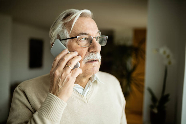 Töprengő öregember néz félre, miközben otthon telefonál..  - Fotó, kép