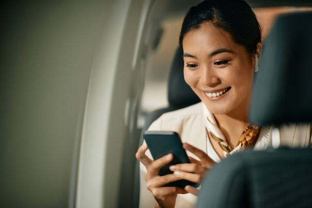 Happy Asian female CEO SMS SMS na mobilním telefonu sedí na zadním sedadle auta. - Fotografie, Obrázek