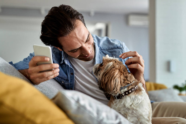Young happy man using mobile phone and having fun with his dog in the living room.  - Foto, imagen