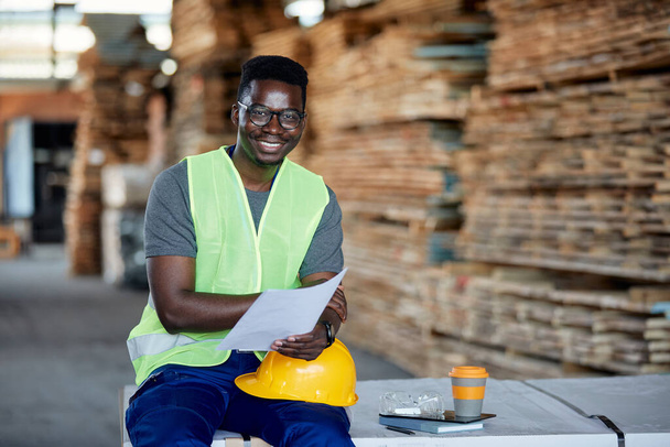 Happy black warehouse worker analyzing plans at lumber distribution department and looking at camera. - 写真・画像