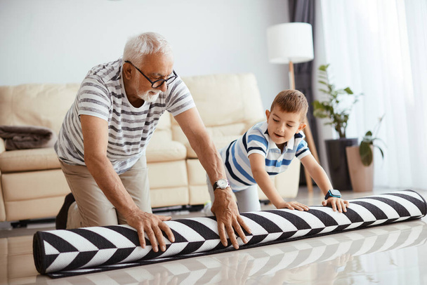Senior man unrolling the carpet with help of his small grandson at home. - Foto, Imagem