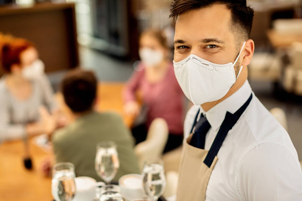 Young happy waiter wearing protective face mask while serving guests and working during COVID-19 epidemic.  - Photo, Image