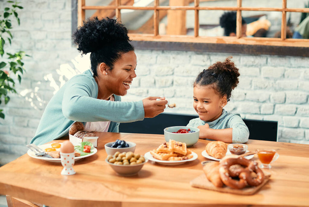 African American mother having fun with her daughter while feeding her during breakfast at home.  - Foto, afbeelding