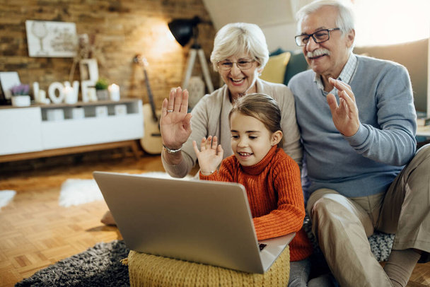 Щаслива маленька дівчинка і її бабуся і дідусь вітають когось під час відеочат на ноутбуці вдома
.  - Фото, зображення