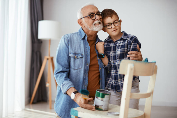 Loving senior man embracing his grandson while spending time with him and painting furniture together at home. - Zdjęcie, obraz
