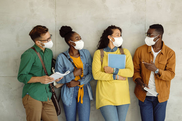 Small group of university students wearing protective face masks while talking by the wall in a hallway.  - Foto, imagen